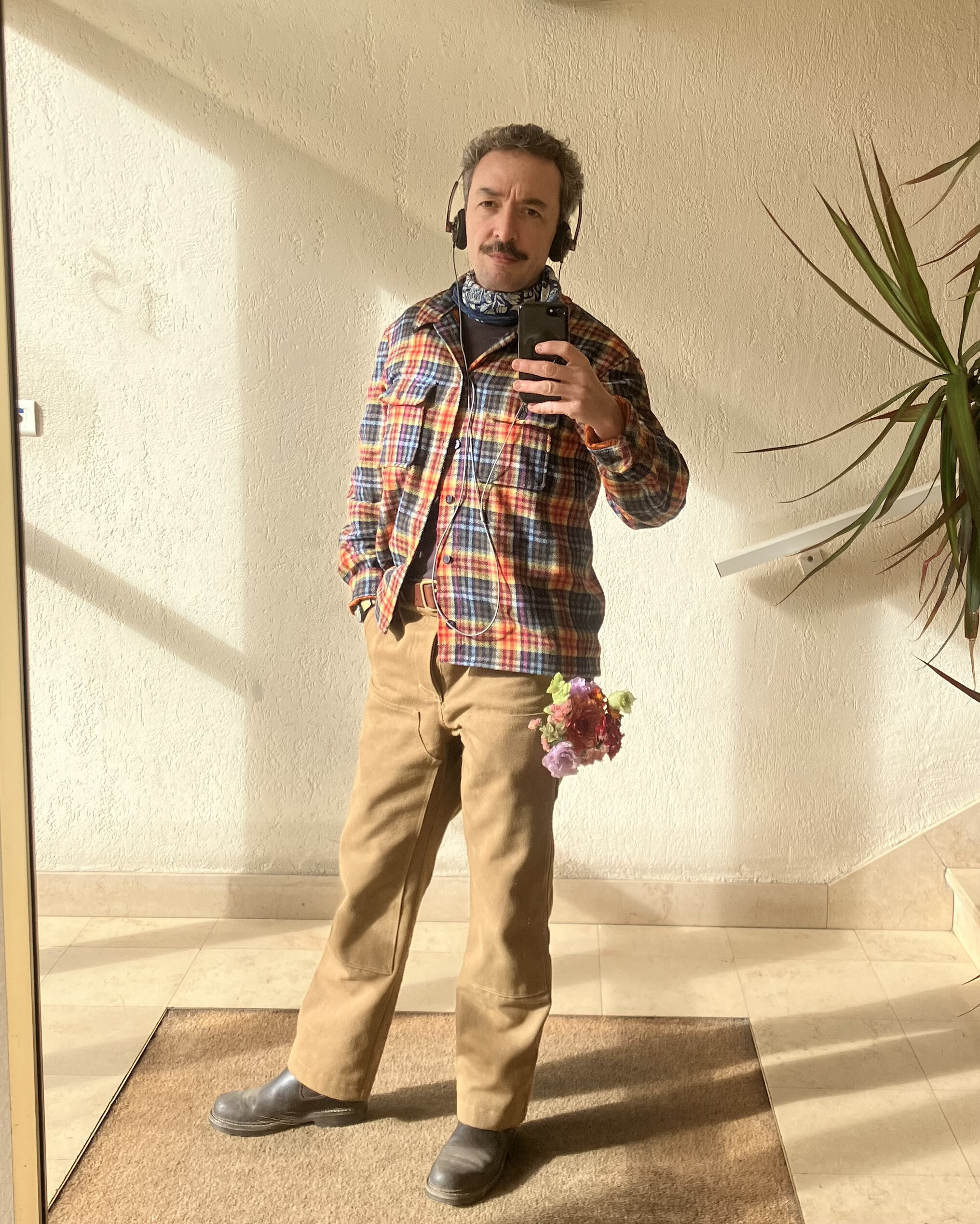 photo en pied du photographe, dans une pièce illuminée par le soleil. il porte une chemise en laine à motif de tartan multicolore qui lui va bien, et un pantalon de travail brun clair. dans la poche de côté de son pantalon il a un petit bouquet de fleurs dont les couleurs peuvent rappeler un peu celles de la chemise