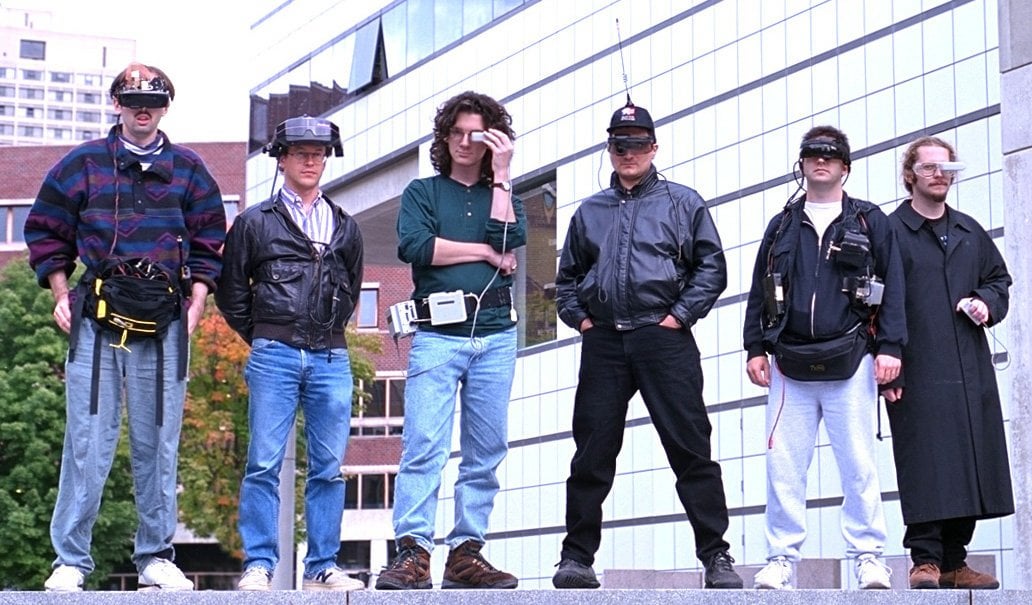 photo de six hommes blancs tous habillés de manière différente des années 90 et portant de la technologie portable et lunettes de réalité augmentée, ils prennent des poses cool mais ont l’air de geeks